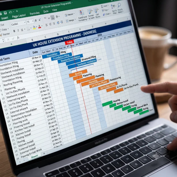 Construction Gantt chart spreadsheet open on a desk beside a builder's programme schedule